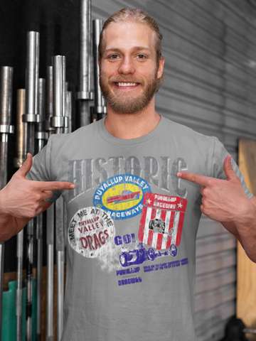 A smiling man with a beard, wearing a grey t-shirt that has "HISTORIC" and various designs related to "Puyallup Valley Raceway" printed on it. He is pointing at the designs on his shirt with both hands. In the background, there are vertical metal bars or pipes against a gray wall.
